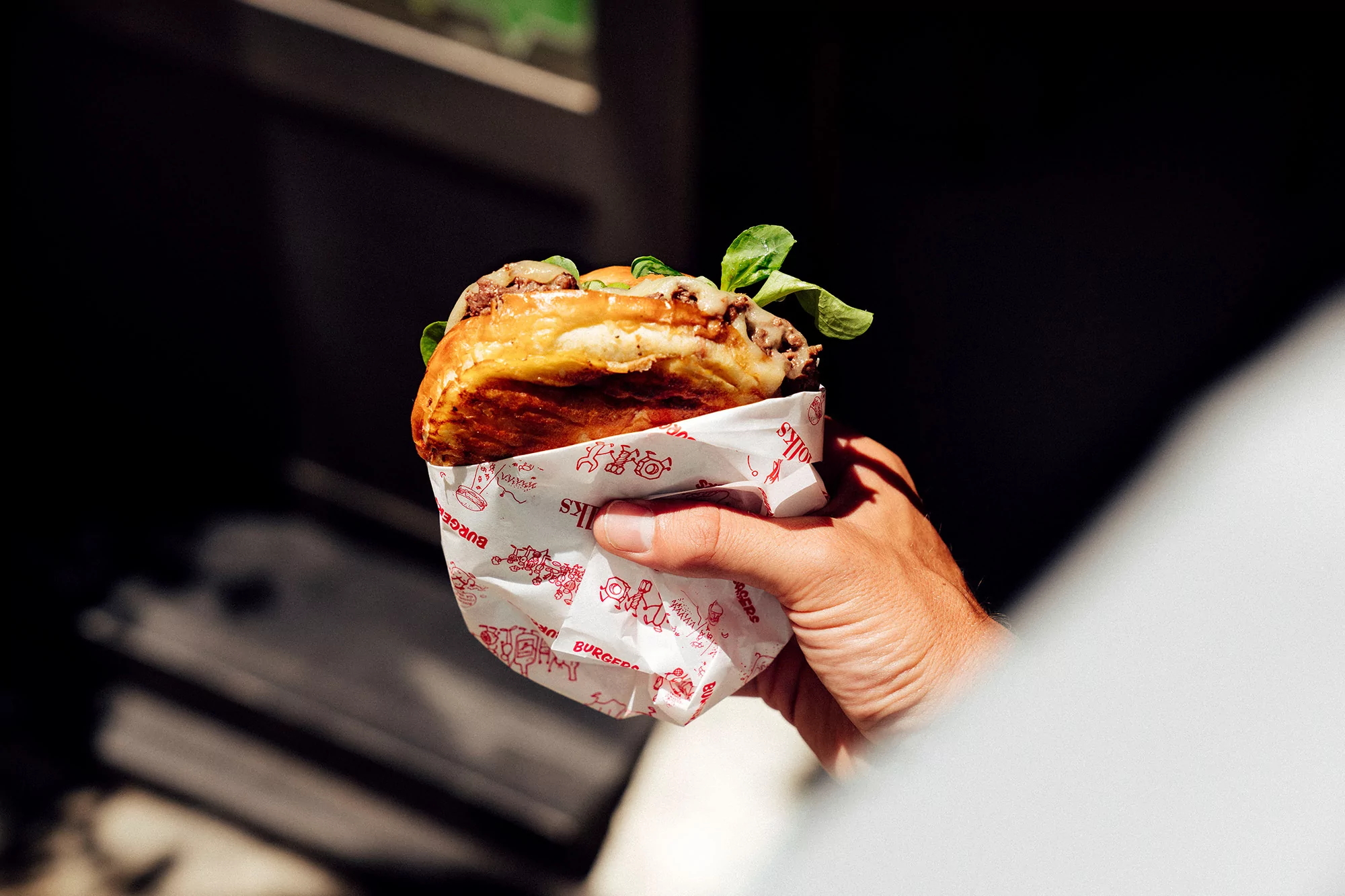 A hand holds a burger wrapped in branded paper, bathed in natural light, showcasing a food product and person in an outdoor setting.