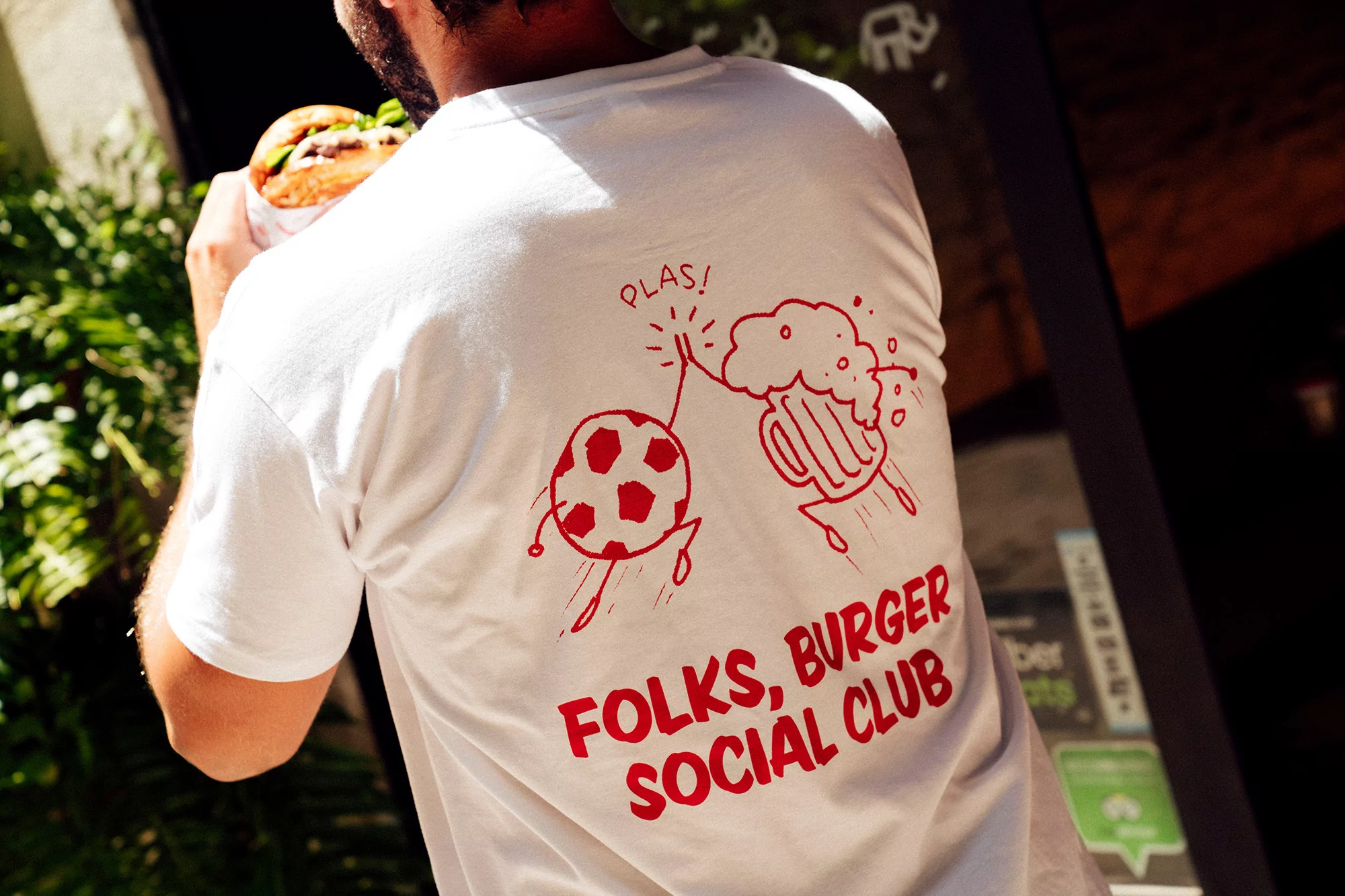 Person in white t-shirt with cartoon soccer ball & beer illustration, holding burger. Burger Social Club text. Outdoors, bright light.