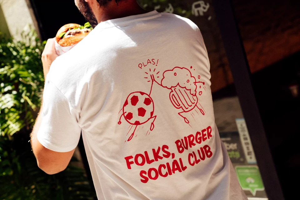 Person in white t-shirt with cartoon soccer ball & beer illustration, holding burger. Burger Social Club text. Outdoors, bright light.