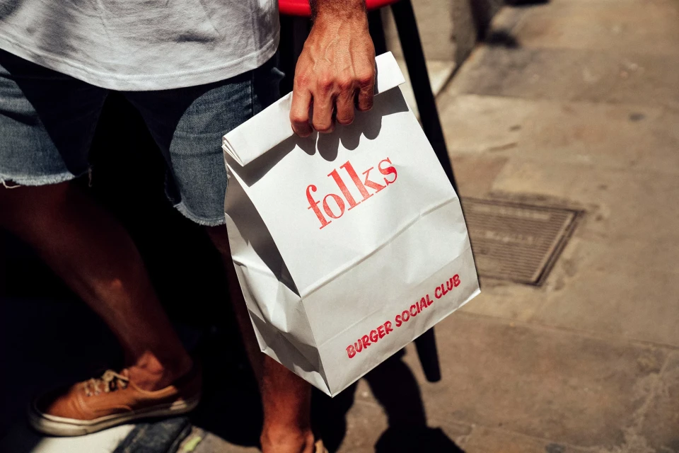 Person holds a white paper bag with 'folks BURGER SOCIAL CLUB' printed in red, wearing denim shorts and tan shoes, outdoors.
