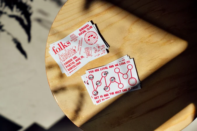 Overhead shot of Folks Burgers Social Club cards on a wooden table with dappled sunlight, promoting loyalty program. Burger restaurant vibe.