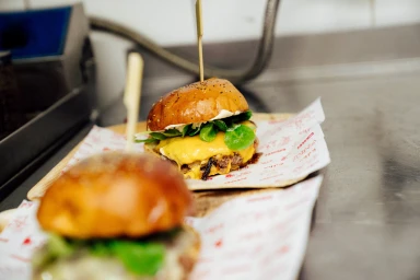 Two gourmet cheeseburgers served on branded paper liners on a metallic commercial kitchen counter