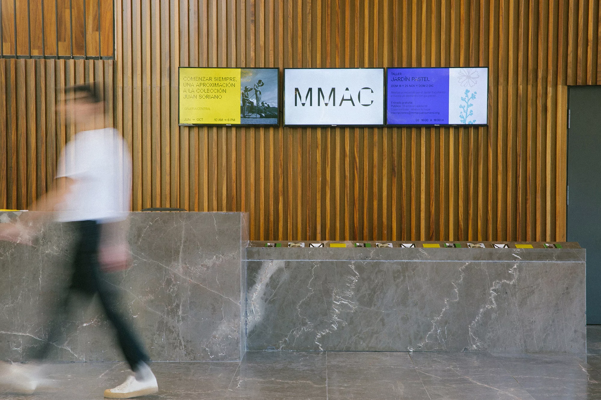 Blurred figure walks past a modern grey stone reception desk, with wooden slat wall and exhibition posters in the background.