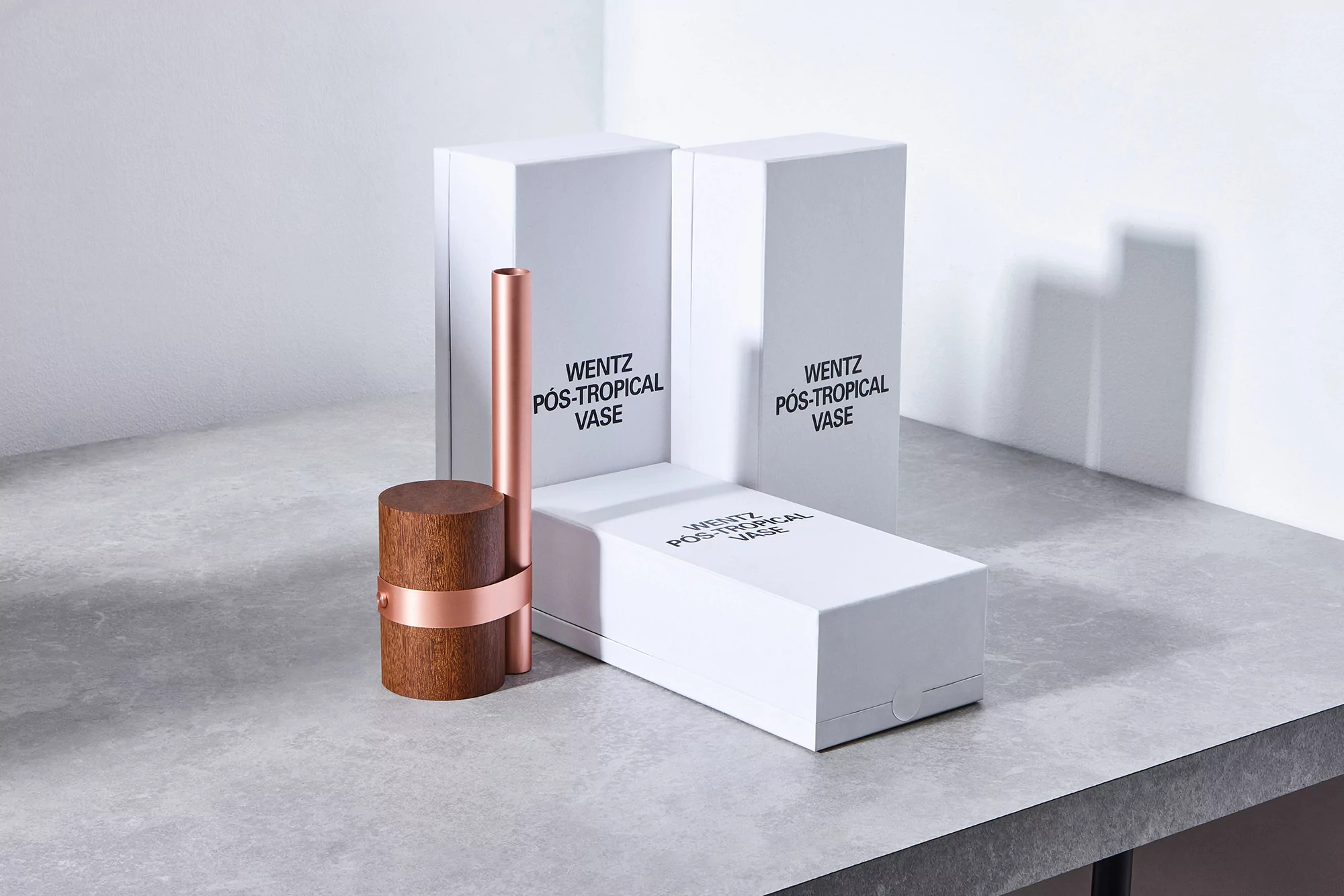 Minimalist vase with wooden base and copper stem displayed alongside sleek white packaging on a grey surface, creating a modern aesthetic.
