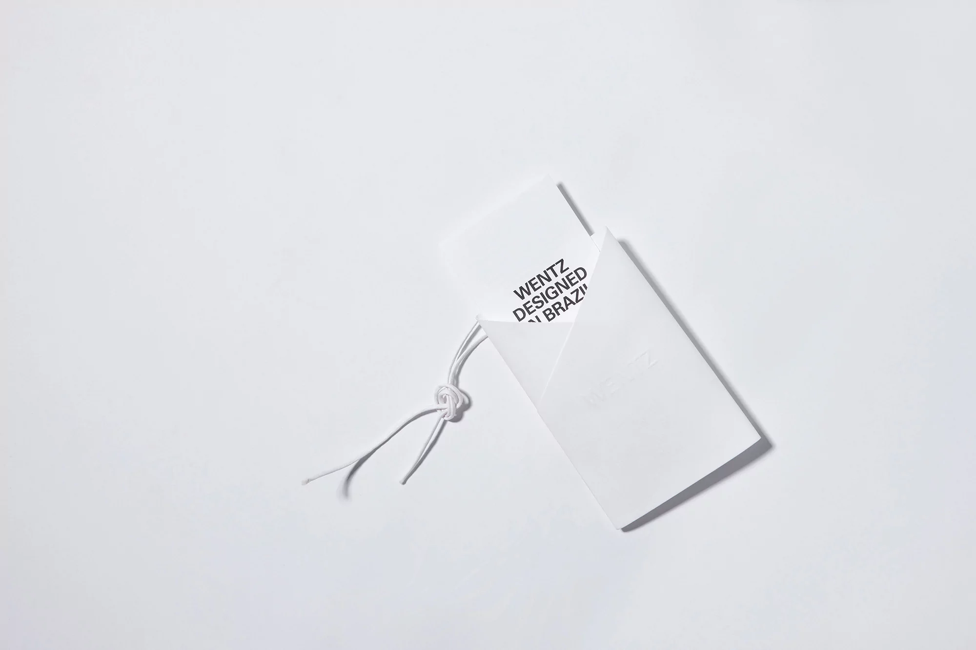 Close-up studio shot of a white envelope with WENTZ DESIGNED IN BRAZIL printed on a white background, featuring a white string.