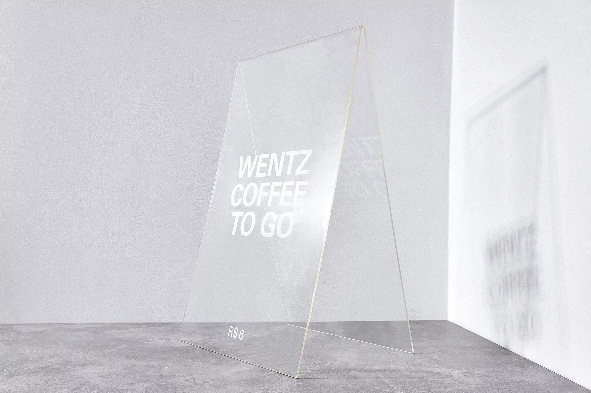 An A-frame sign with translucent surfaces features the text 'Wentz Coffee To Go', displayed on a gray floor against a white wall.