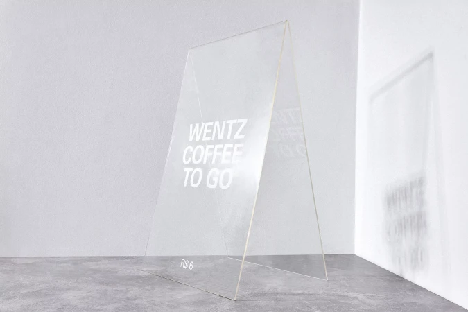An A-frame sign with translucent surfaces features the text 'Wentz Coffee To Go', displayed on a gray floor against a white wall.