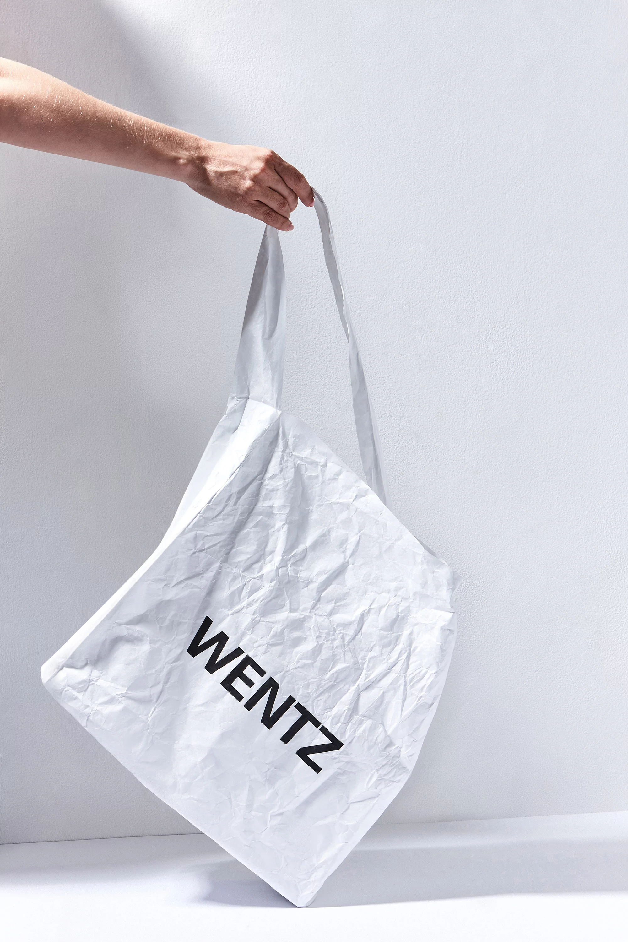 Hand holds a white tote bag with the word "WENTZ" in black letters, against a white wall with natural light and shadows.