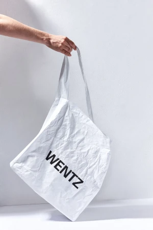 Hand holds a white tote bag with the word "WENTZ" in black letters, against a white wall with natural light and shadows.