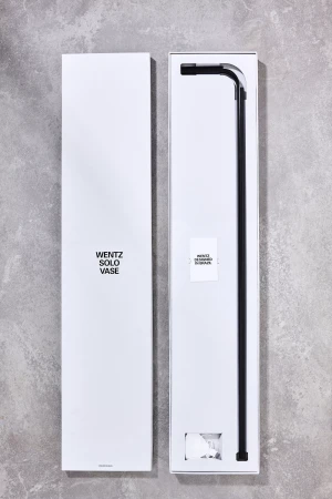 Wentz Solo Vase in open packaging showing a long black rod with a 90-degree bend at the top, minimalist product presentation.