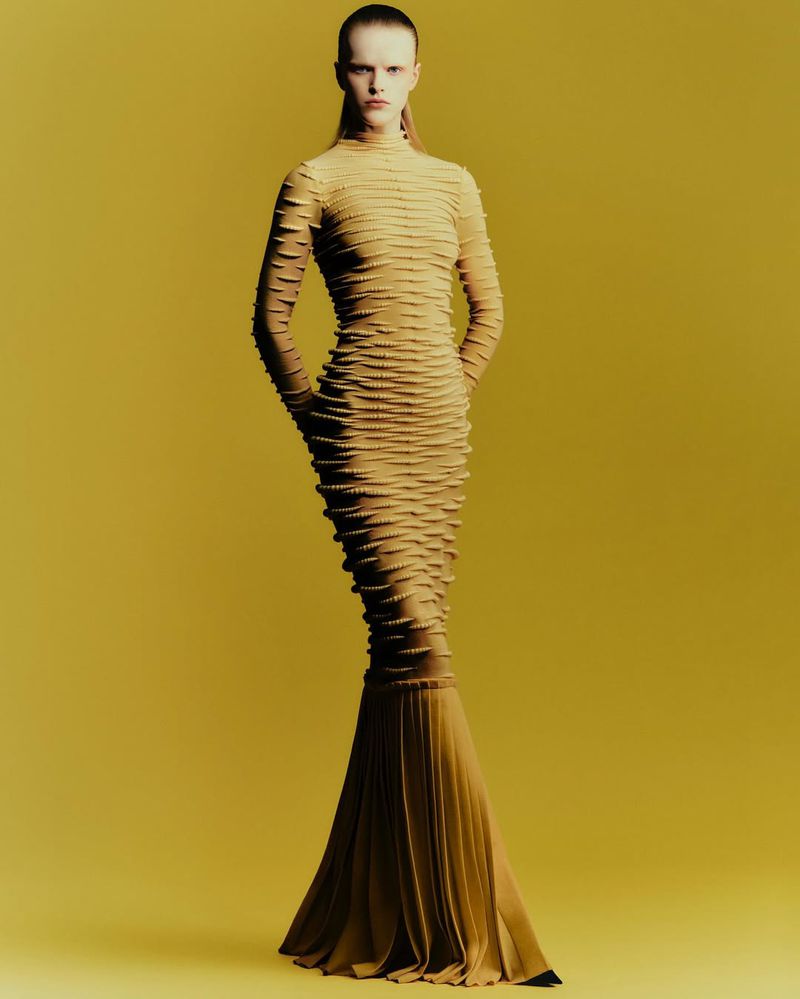 A single fair-skinned adult woman is standing in a photo studio modeling a long, tan textured dress with hands clasped against a yellow backdrop.