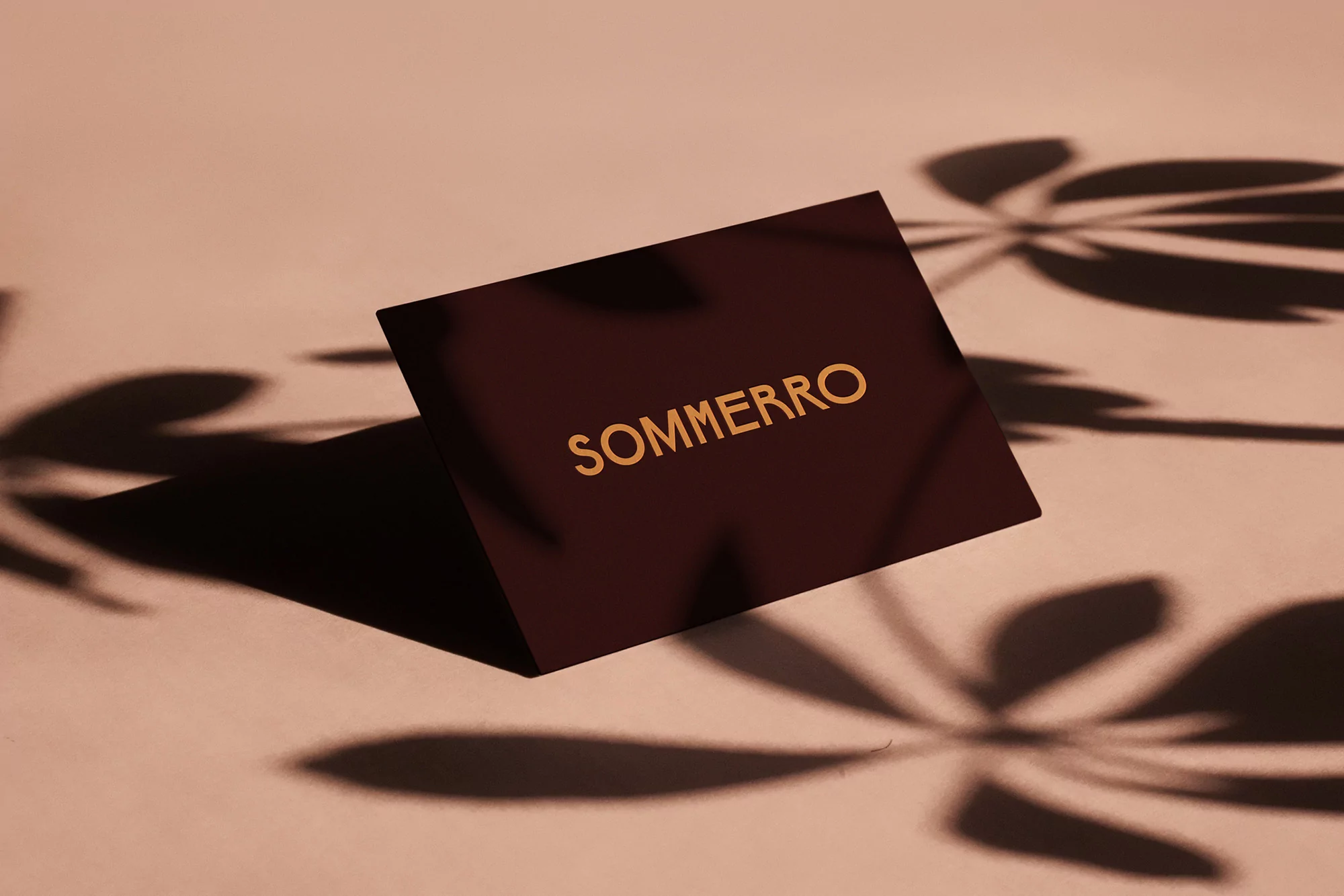 Promotional shot featuring a maroon business card with the word SOMMERRO in gold lettering against a light pink background with leaf shadows.