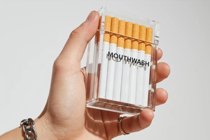 Hand holds clear acrylic box filled with white cigarettes labeled 'MOUTHWASH' against a plain background, showcasing a conceptual design.