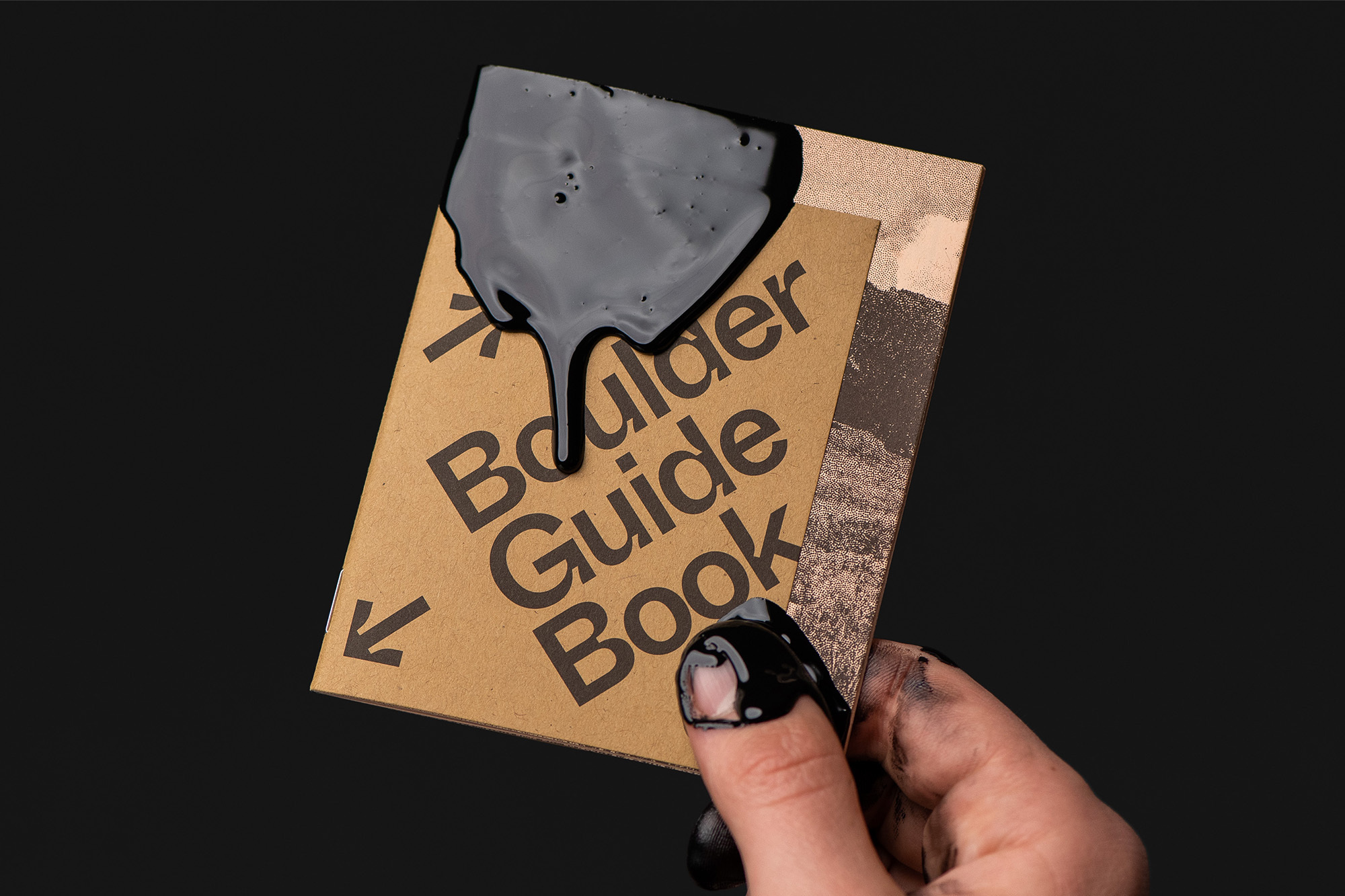 Close-up of hand holding a 'Boulder Guide Book' covered in black ink, against a dark background. Features abstract design elements.