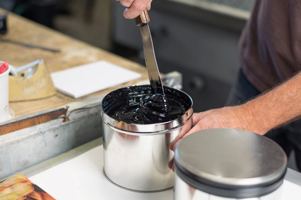Close-up of artist's hands using a knife to mix black ink in a silver tin can at a workbench in an indoor studio.