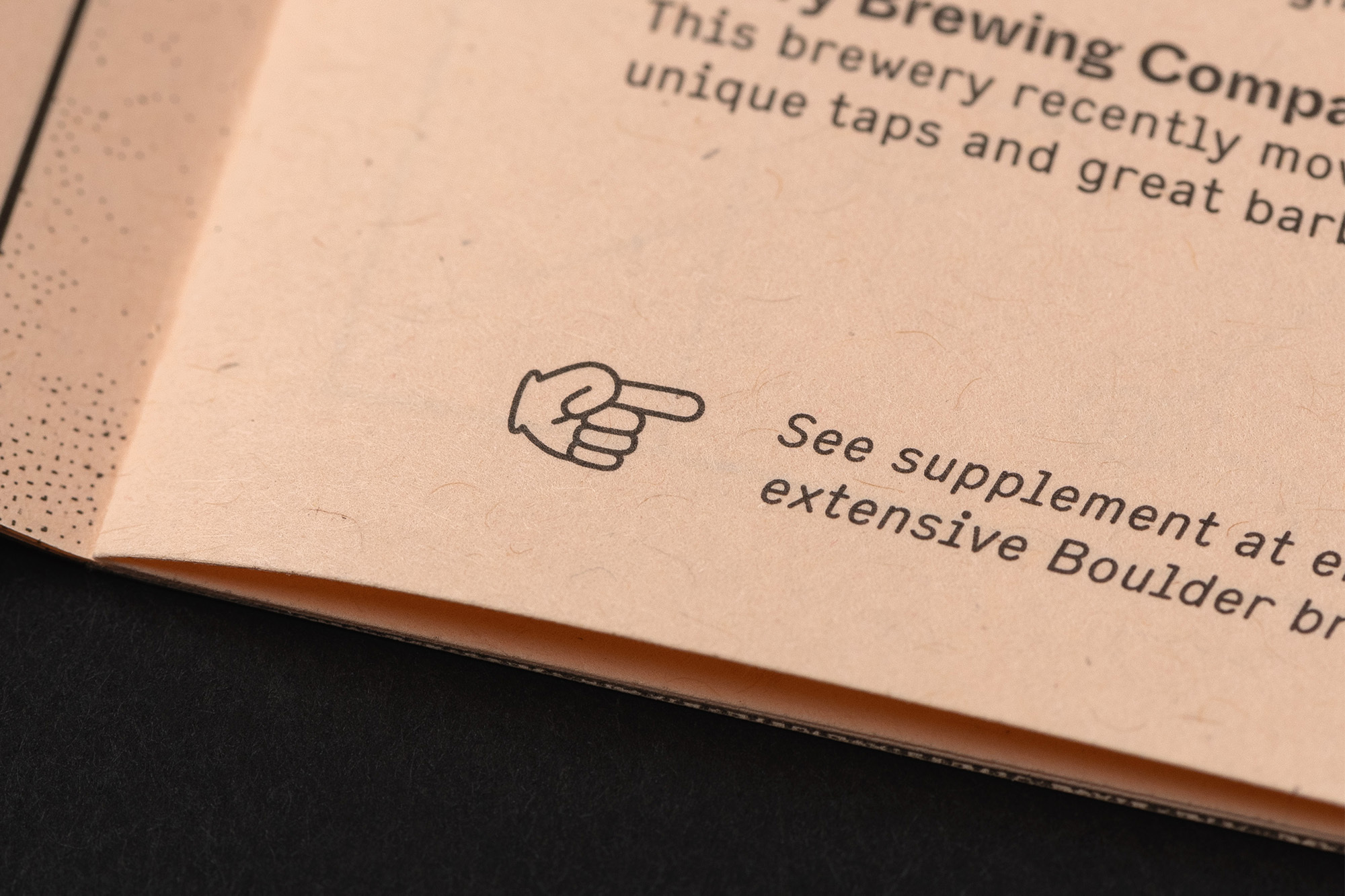 Close-up of an open booklet on a black surface, revealing text about brewing and a pointing hand icon directing the viewer.