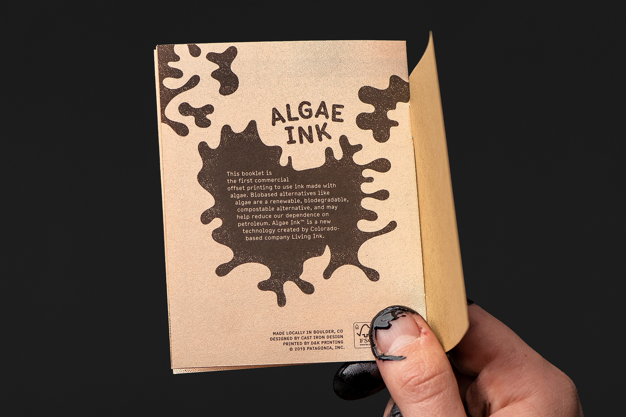Open booklet on kraft paper with algae ink text and design held by a hand with ink-stained fingernails, presented on a black background.