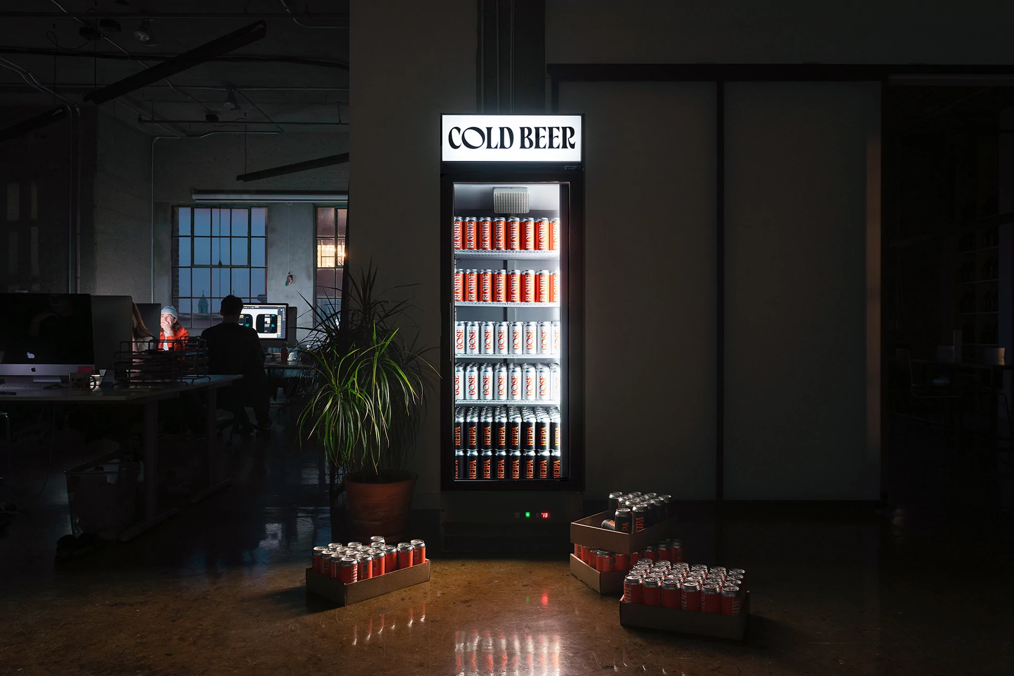 Illuminated beer refrigerator in an office setting, stocked with cans. Crates of canned beverages on the floor create an inviting ambiance.