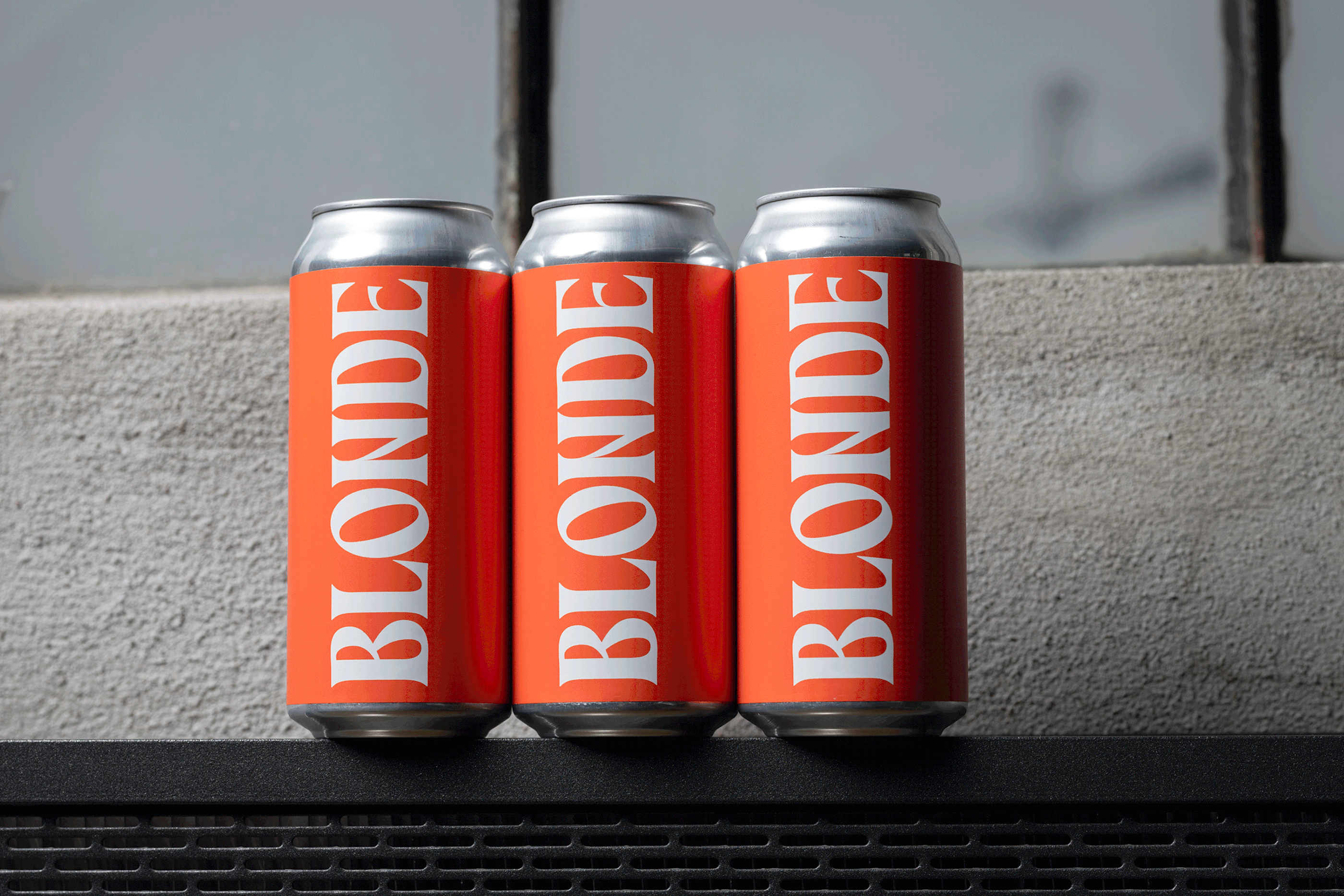 Three aluminum cans with orange labels displaying the word 'BLONDE' in white, standing on a black surface with a blurred background.