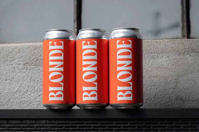 Three aluminum cans with orange labels displaying the word 'BLONDE' in white, standing on a black surface with a blurred background.
