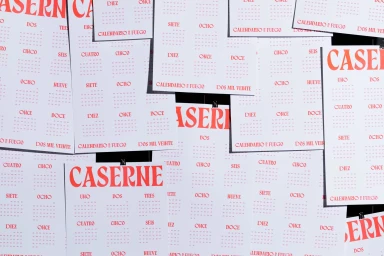 Multiple white printed calendar sheets with red typography featuring CASERNE displayed against a dark surface