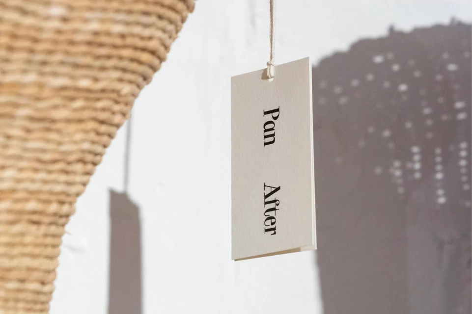 Close-up of a straw basket with a tag hanging from a string in front of a white wall, creating light and shadow patterns.