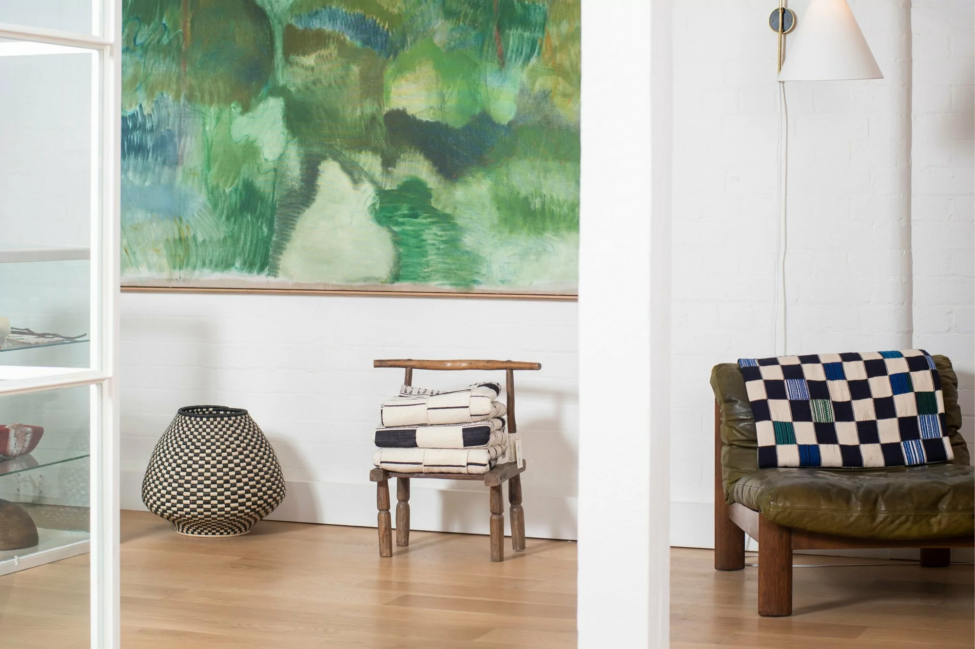 Living room interior with white walls, a painting, a black and white woven basket, chair with towels, and leather armchair with a blanket.
