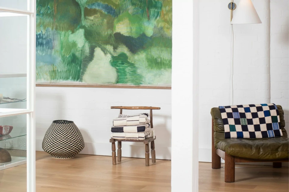 Living room interior with white walls, a painting, a black and white woven basket, chair with towels, and leather armchair with a blanket.