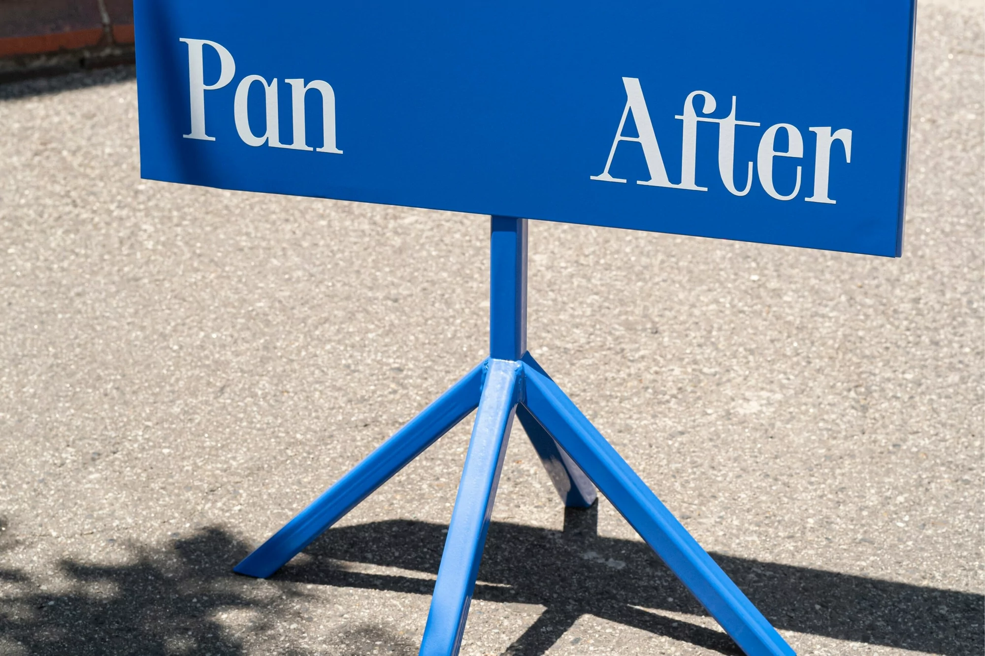 Blue sign on a metal tripod with white text reading 'Pan' and 'After', set on a concrete surface with shadows visible.