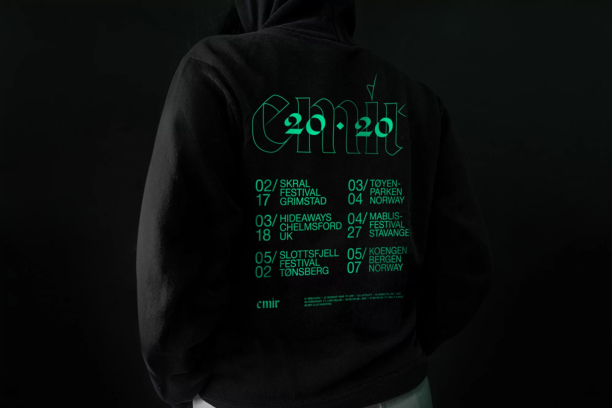 Back view of a person wearing a black hoodie with a neon green graphic of tour dates and locations, set against a dark background.