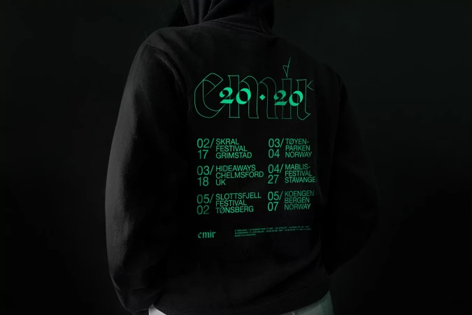 Back view of a person wearing a black hoodie with a neon green graphic of tour dates and locations, set against a dark background.