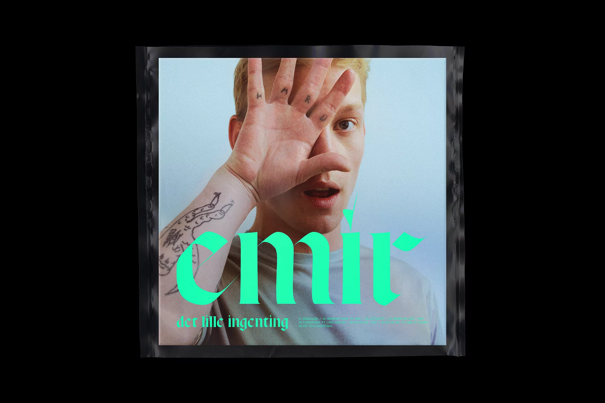 Album cover featuring a young adult with blond hair obscuring one eye with his hand, mint green graphic text and arm tattoo.