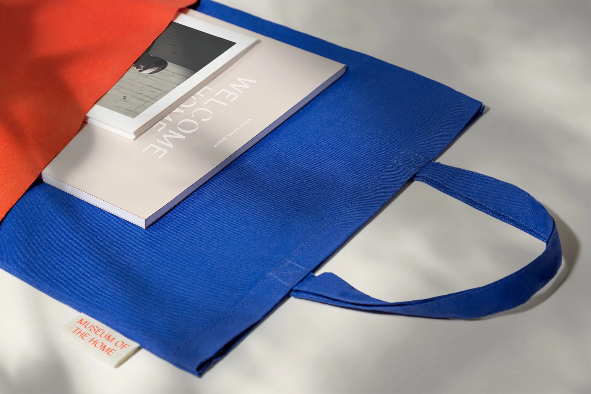 Open blue and orange tote bags display an open invitation to 'Welcome Home,' with a brochure featuring a photograph.