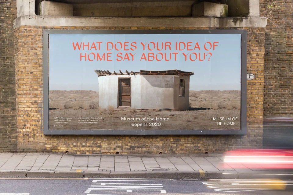 Billboard on a brick wall advertises the Museum of the Home with an image of a small building in a deserted landscape under a blue sky.