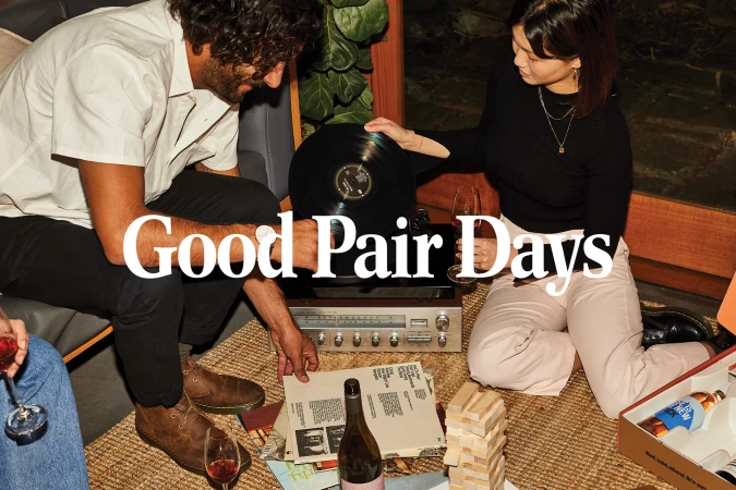 Two people gather around a turntable in a cozy indoor setting, enjoying records, wine, and a block-stacking game, showcasing a relaxed social scene.