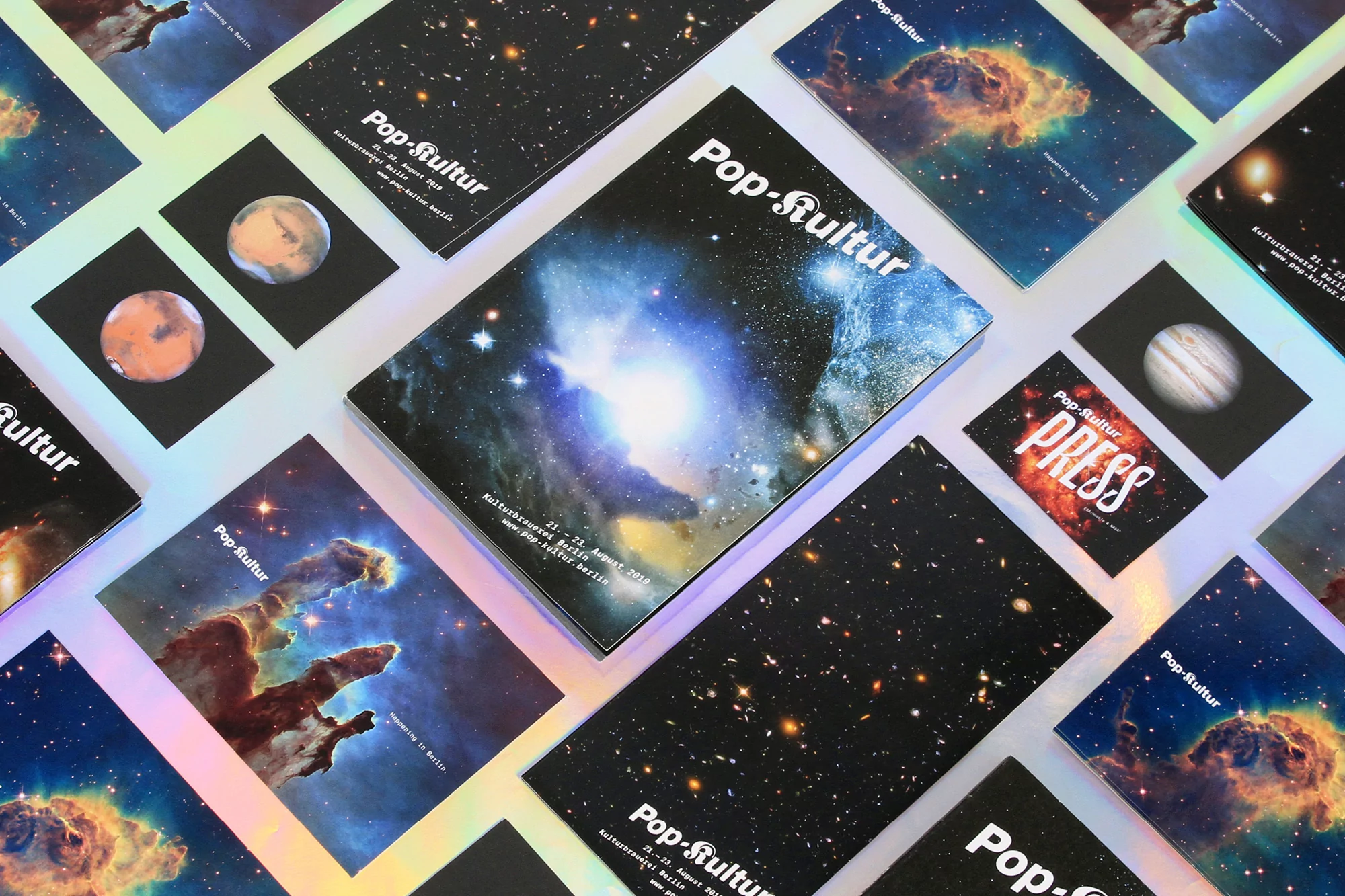 Stack of Pop-Kultur magazines and cards with galaxy and planet imagery on a holographic surface, laid out in a visually engaging pattern.