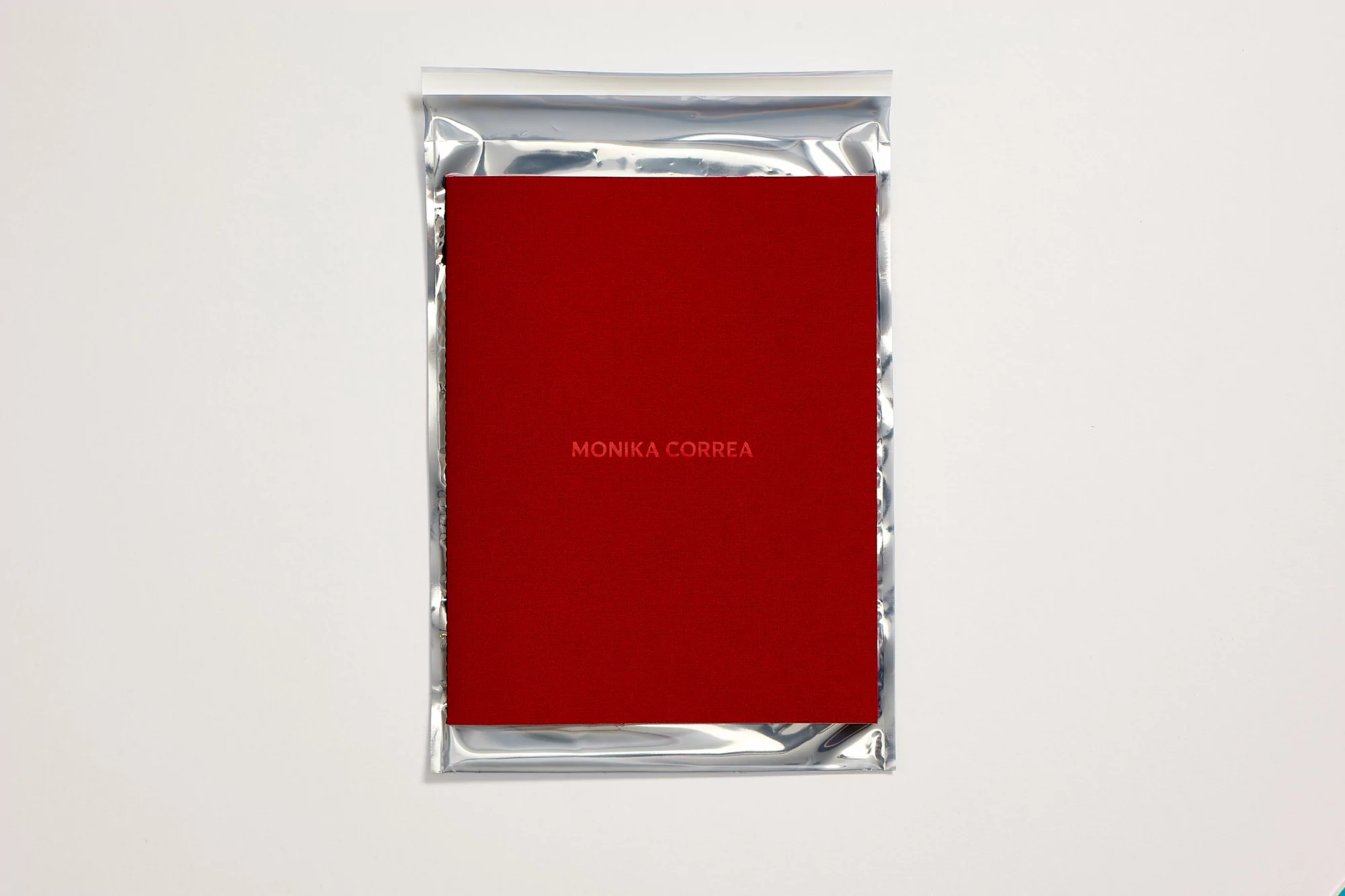 Studio shot of a red square book with 'MONIKA CORREA' text, encased in a shiny silver packet, set against a stark white background.