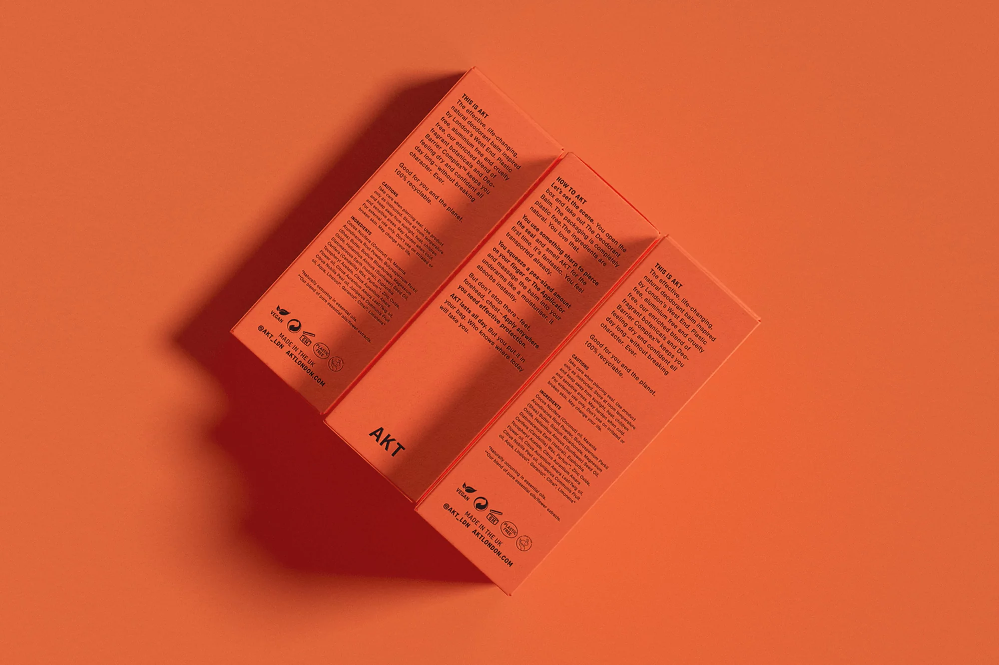 Three orange AKT deodorant boxes are stacked against a matching background. Text and logos are printed on the box sides.
