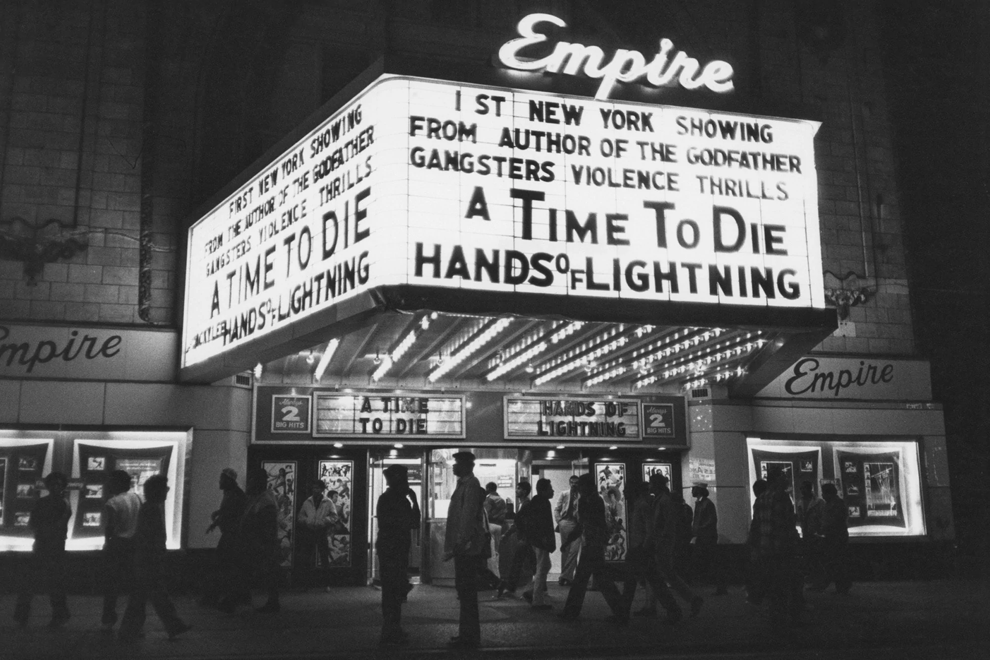 Vintage black and white image of a large group of people gathered at night in front of the Empire theater with film titles on the marquee.