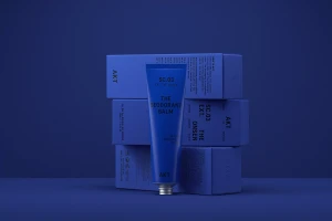 Blue deodorant tube standing vertically in front of stacked blue packaging boxes against a dark blue monochromatic background.