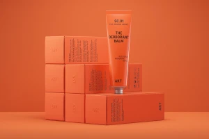 Deodorant balm tube standing upright on stacked orange product boxes against a matching orange background