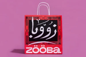 Red paper shopping bag with white handles featuring Arabic calligraphy and the brand name Zooba against a pink background