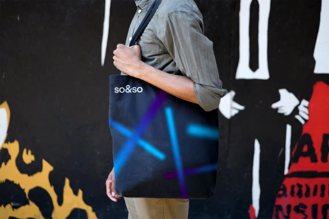 Adult male carries a black tote bag with bright blue and purple neon-style graphics in front of a colorful mural wall outdoors.