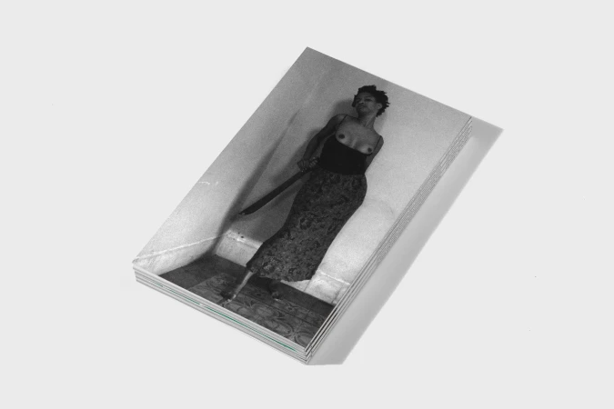 Monochrome photograph print shows a topless African American woman holding a machete, posing in a corner on a patterned floor.