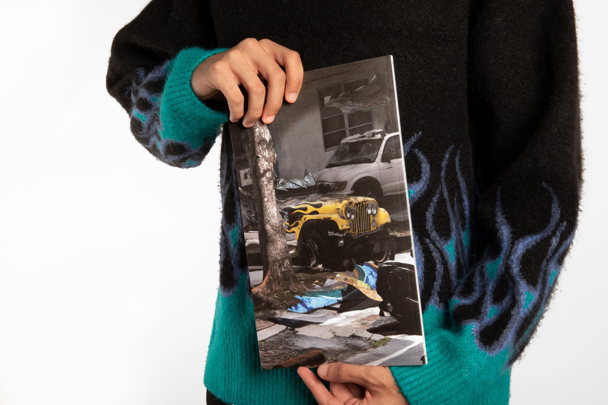 Person holding a book cover featuring a yellow vehicle in a street scene, contrasting with the individual's black and green flame sweater.