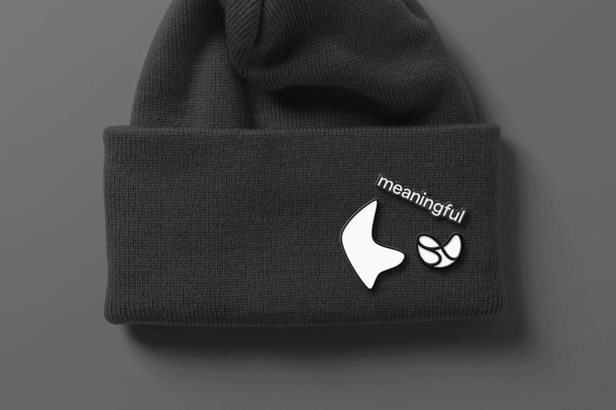 Close-up of a dark gray knit beanie, adorned with white abstract designs and the text 'meaningful' against a neutral gray background.