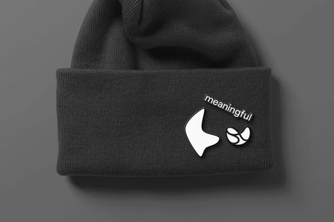 Close-up of a dark gray knit beanie, adorned with white abstract designs and the text 'meaningful' against a neutral gray background.