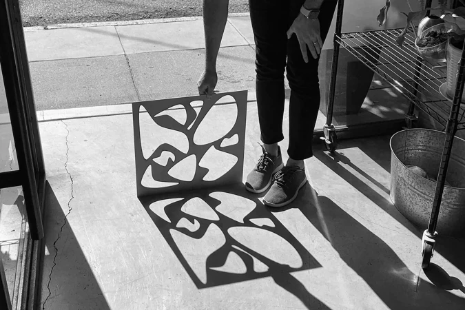 Person holds a patterned panel creating shadows on the floor indoors, featuring a metal shelving unit and gray sneakers in black and white.