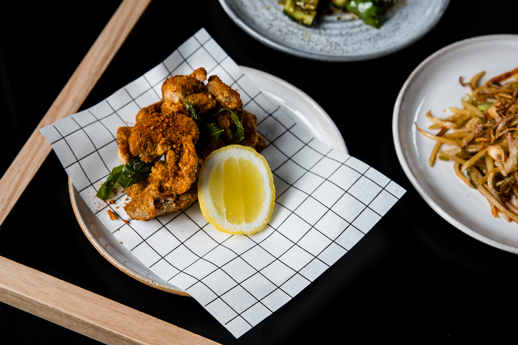 Overhead shot of orange-toned fried food with lemon on a gridded paper-lined plate, and noodles and vegetables in the background.