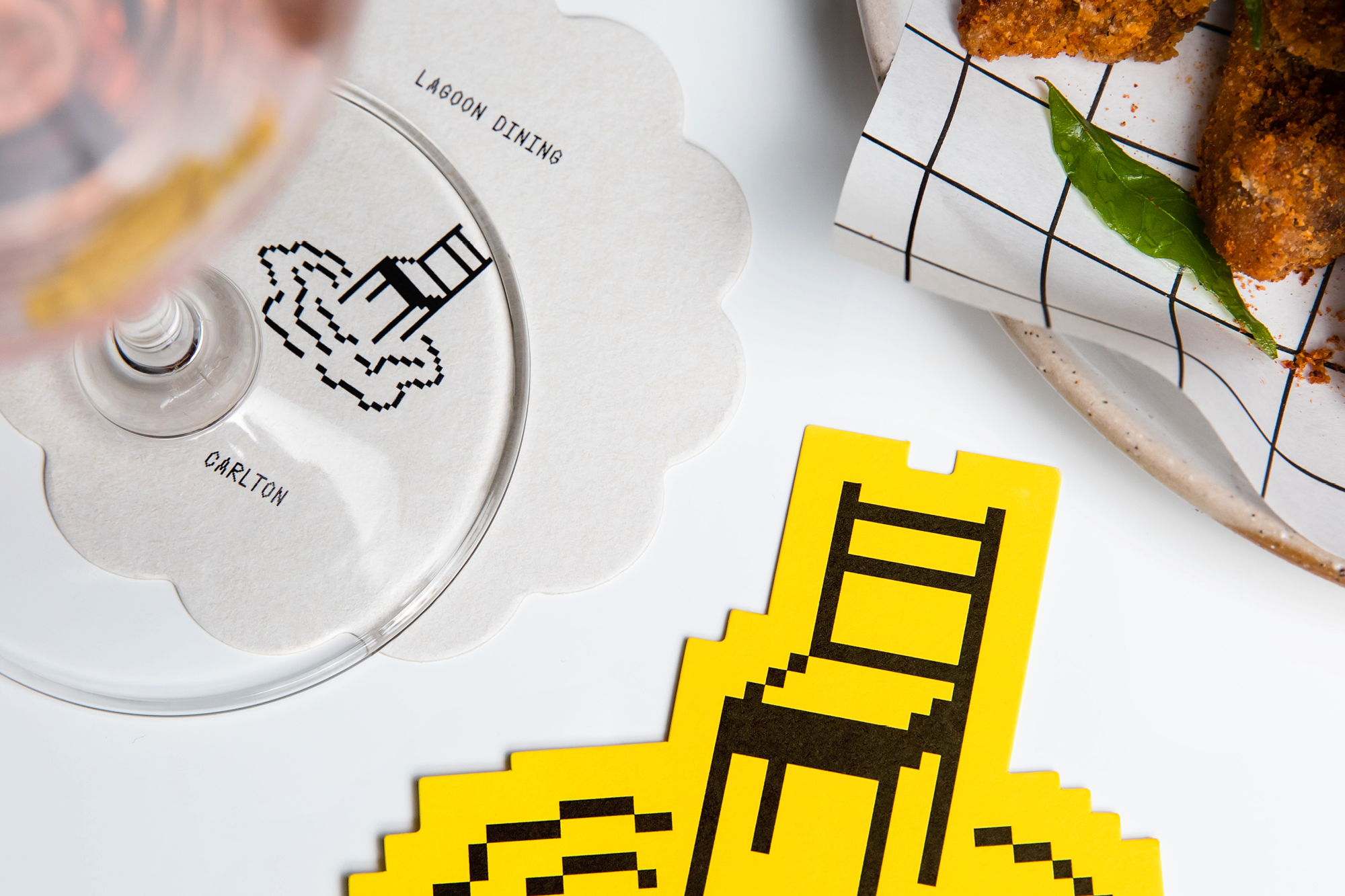 Overhead shot of a glass and coasters with pixelated chair designs, next to a plate of food on a white table, commercial setting.
